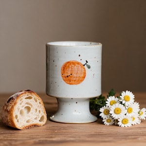 May include: A light blue ceramic pedestal cup with an orange design. The cup has brown speckles and a wide base. A slice of bread and a small bouquet of daisies are next to the cup on a wooden surface.