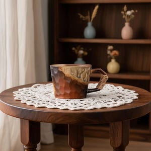 May include: A textured ceramic mug with a brown and tan gradient sits on a round wooden table. The mug's handle is wrapped with a woven material. A white lace doily is beneath the mug. The background includes a wooden shelf with small vases.