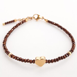 Könnte beinhalten: Ein zartes Armband mit kleinen, facettierten dunkelbraunen Perlen an einer goldfarbenen Kette. Ein glatter, polierter goldfarbener Herzanhänger ist mittig platziert. Das Armband verfügt über einen goldfarbenen Karabinerverschluss und eine Verlängerung.