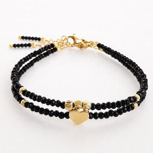 May include: A double-strand bracelet featuring small, faceted black beads and gold-toned accents. The design includes a central gold-toned heart charm flanked by three small gold-toned beads, finished with a gold-toned lobster claw clasp.