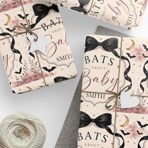 May include: Wrapping paper featuring a cream background with a repeating pattern of black bows, bats, gold crescent moons, stars, and pink clouds. The text reads BATS ABOUT BABY SMITH. Each package is tied with brown twine and a white star tag.