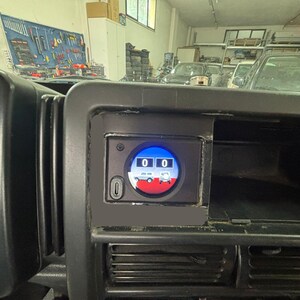 Op de afbeelding: Close-up van een voertuigdashboard met een digitale display met de nummers "00". De display heeft een rood, wit en blauw grafisch ontwerp van een voertuig. Het dashboard is zwart, met een snelheidsmeter zichtbaar aan de linkerkant.