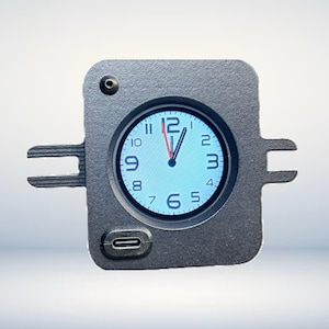 May include: A square, gray watch case with a digital clock face. The clock shows the time as 12:01. The case has two protruding arms on either side and a USB-C port at the bottom. A small circular button is visible at the top.