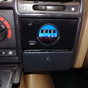 Könnte beinhalten: Schwarzes Auto-Armaturenbrett mit digitaler Anzeige, die die Zahl "00" anzeigt. Das Display hat einen blauen Hintergrund mit kleinen Autosilhouetten. Ein USB-Anschluss ist unter dem Display sichtbar. Das Armaturenbrett enthält auch Klimaanlagenregler und einen Zigarettenanzünder.
