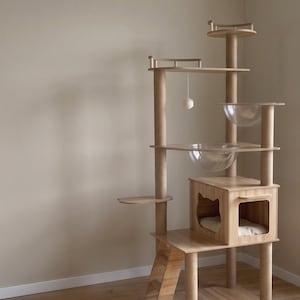 Könnte beinhalten: Ein mehrstöckiger Katzenbaum aus hellem Holz mit mehreren mit Sisal umwickelten Kratzstämmen, zwei durchsichtigen Acrylschalen als Liegeflächen, einer kleinen geschlossenen Höhle mit katzenförmigem Ausschnitt, einer Rampe und einem Spielball.