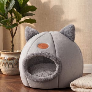Pode incluir: Uma cama cinza para gatos em forma de cúpula com orelhas de gato. A cama tem um interior macio e felpudo e uma borda de pele sintética ao redor da entrada. Um pequeno remendo de couro marrom está na frente. Um cobertor dobrado está ao lado da cama.
