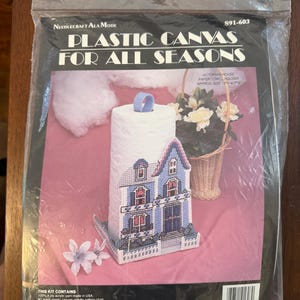 May include: A Needlecraft Ala Mode plastic canvas kit for a Victorian house paper towel holder. The packaging features the text Plastic Canvas For All Seasons and S91-603. The kit includes a pattern for a blue and white house-shaped holder.