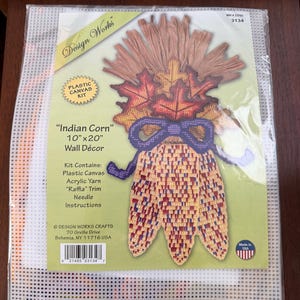 Könnte beinhalten: Ein Design Works Kunststoff-Stickset für eine Indian Corn Wanddekoration mit den Maßen 25,4 cm mal 50,8 cm. Das Set enthält Kunststoff-Stramin, Acrylgarn, Bast-Verzierung, eine Nadel und eine Anleitung mit einer Abbildung von Mais und Blättern.