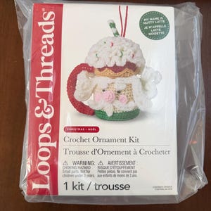 May include: A Loops & Threads crochet ornament kit in a plastic bag. The packaging features a white box with a red side panel, displaying a crocheted mug ornament with a Santa face, whipped cream top, and red handle. Text includes My Name Is Nutty Latte.