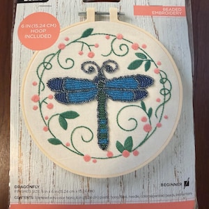 May include: A Bucilla beaded embroidery kit featuring a dragonfly design on ecru fabric within a 6-inch plastic hoop. The kit includes stamped fabric, floss, a needle, and beads. The packaging displays the finished dragonfly with blue wings.