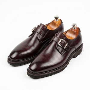 May include: A pair of dark brown leather monk strap shoes featuring a single silver-toned metal buckle closure, a rounded toe, and a thick, lugged rubber sole. Each shoe contains a wooden shoe tree insert with a polished metal knob handle.