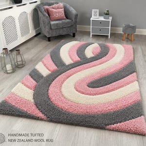 May include: A rectangular handmade tufted rug featuring a high-pile texture with a repeating curved pattern in shades of soft pink, cream, and dark gray. The design consists of thick, layered, and sweeping bands that create a sense of depth.