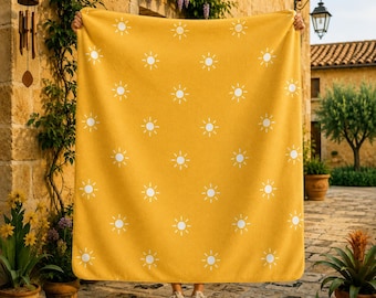 May include: A mustard yellow blanket featuring a repeating pattern of white sun icons. The blanket is rectangular with finished edges and is held up to display the full design, which consists of small, stylized suns with radiating rays.