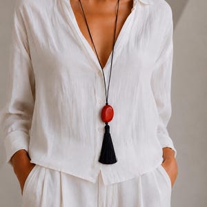 May include: A long necklace featuring a thin black cord, a smooth oval-shaped red stone pendant, and a black tassel hanging below the stone. The necklace is worn over a white button-down shirt with long sleeves and matching white trousers.