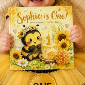 May include: A square book titled Sophie is One! Sweet as Honey, One Year Old, featuring a cartoon bee, a birthday cake with a number one candle, and sunflowers. The book cover has a yellow and white color scheme with honeycomb patterns.