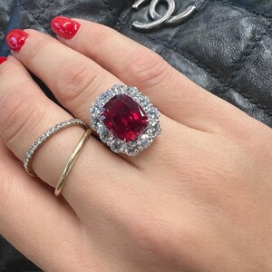 May include: A ring featuring a large, cushion-cut deep red gemstone surrounded by a halo of small, clear, round-cut stones. Two additional thin bands are worn on the same finger, one plain gold and one set with tiny clear stones.