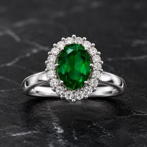 Op de afbeelding: Een zilverkleurige ring met een grote, ovaal geslepen, diepgroene middensteen. De centrale edelsteen is omringd door een halo van kleine, ronde, heldere, fonkelende steentjes in een geschulpte zetting, gemonteerd op een gladde, gepolijste band.