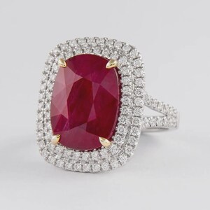 Könnte beinhalten: Ein Ring mit einem großen, kissenförmigen, tiefroten Rubin, der in Gelbgoldfassungen sitzt. Der Mittelstein ist von einem doppelten Halo aus kleinen, runden, im Brillantschliff geschliffenen weißen Diamanten in Weißmetall umgeben.