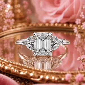 May include: A silver-toned ring featuring a large, rectangular emerald-cut center stone held by four prongs. The band is accented on each side by two marquise-cut stones arranged in a leaf-like pattern, creating a symmetrical and detailed design.
