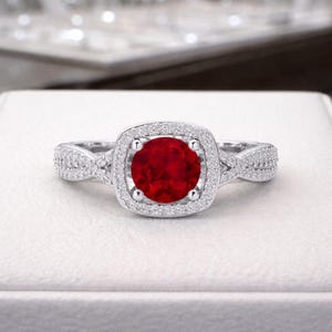 Puede incluir: Un anillo de tono plateado con una piedra central redonda de color rojo vibrante, engastada en un halo cuadrado de pequeñas piedras transparentes en pavé. La banda se divide en dos hilos entrelazados con pavé que se unen en el centro.