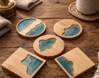 May include: A set of five coasters made from light-colored wood and blue resin. Three coasters are circular and two are square. Each piece features a layered, topographical design that mimics the appearance of deep blue water and sandy shores.