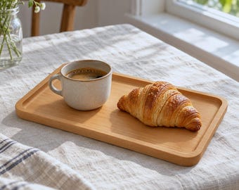 May include: A rectangular light-colored wooden serving tray holding a speckled off-white ceramic mug filled with dark coffee and a golden-brown flaky croissant. The tray has rounded corners and a smooth finish, providing a stable base.
