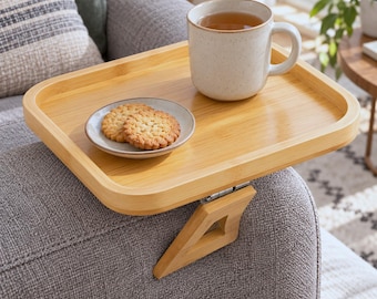 May include: A rectangular bamboo sofa arm tray with a hinged clamp mechanism. The tray holds a speckled off-white ceramic mug filled with tea and a small round plate containing two round golden-brown cookies with a textured surface.