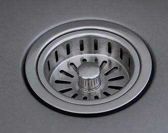 May include: A circular stainless steel kitchen sink drain strainer with a central knob and a series of vertical and radial slots for water drainage, set flush into a matte gray sink basin surface with a visible black rubber gasket seal.