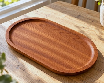 May include: An oval wooden tray with a smooth, polished finish and a visible natural grain pattern. The tray features a shallow, recessed center and a raised, rounded rim, showcasing a warm, medium-brown wood tone throughout the entire piece.