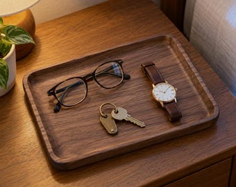 May include: A rectangular dark brown wooden valet tray holding a pair of black-rimmed eyeglasses, a wristwatch with a brown leather strap and a white face, and a set of metal keys with a small brass-colored tag on a key ring.