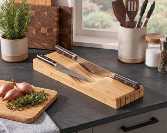 May include: A rectangular bamboo in-drawer knife organizer with multiple horizontal slots. Two stainless steel kitchen knives with dark wooden handles are resting inside the slots. The organizer is placed on a dark countertop.