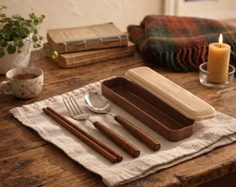 May include: A portable cutlery set featuring a fork, a spoon, and a pair of chopsticks, all with dark brown wooden handles. The set includes a rectangular brown plastic case with a ribbed beige lid, resting on a light-coloured fabric cloth.
