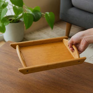 Puede incluir: Una bandeja de servicio rectangular de bambú con un acabado de grano de madera marrón claro natural. La bandeja cuenta con bordes laterales elevados, asas recortadas integradas en los extremos cortos y una base plana para transportar objetos.