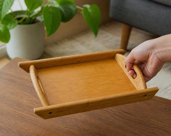 May include: A rectangular bamboo serving tray with a natural light brown wood grain finish. The tray features raised side edges, integrated cutout handles on the shorter ends, and a flat base, designed for carrying or displaying various items.