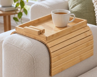 May include: A flexible bamboo sofa arm tray with a flat rectangular surface and a built-in phone stand. The tray features a slatted design that drapes over the armrest, holding a white ceramic mug with vertical ridges filled with tea.
