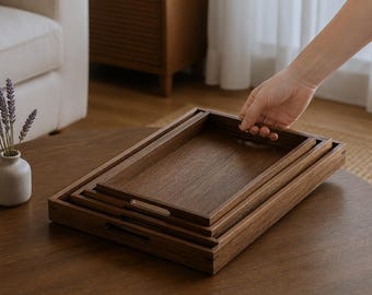 May include: A set of three rectangular nesting trays crafted from dark brown wood. Each tray features a smooth, flat base, raised sides, and integrated cutout handles on the shorter ends. The trays are stacked together, showing a consistent wood grain.