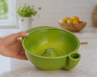 May include: A bright lime green ceramic yarn bowl with a smooth, glossy finish. The bowl features a central raised dome with small holes, a series of small circular openings along the rim, and a single cylindrical side attachment for tools.