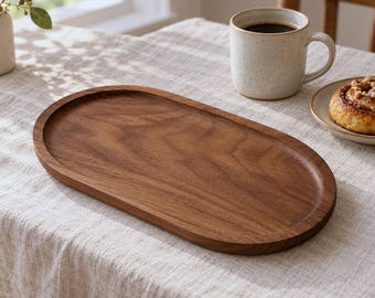 May include: An oval-shaped wooden serving tray with a smooth, polished surface and visible natural wood grain patterns. The tray features a shallow, recessed center and a raised, rounded rim, showcasing a rich, warm brown color.