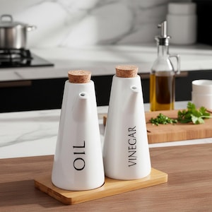 Könnte beinhalten: Zwei weiße Öl- und Essigspender aus Keramik mit Korken, beschriftet mit "OIL" und "VINEGAR". Sie stehen auf einem Bambustablett. Im Hintergrund befindet sich eine klare Glasflasche mit Öl, was auf eine Küchenumgebung hindeutet.
