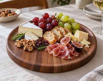 May include: A round wooden serving board holding an assortment of food including red grapes, green grapes, sliced baguette, brie cheese, hard cheese cubes, prosciutto, dried cranberries, walnuts, fresh fig halves, and rosemary sprigs.