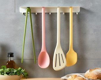 May include: A set of colorful kitchen utensils hanging on a white rack. The set includes green tongs, a pink spoon, a cream-colored slotted spatula, and a yellow spatula. The utensils are hanging against a gray tiled wall.