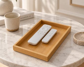 May include: A rectangular wooden tray with a light brown finish, containing two white remote controls. The tray features a flat base and raised edges, providing a simple and functional surface for organizing small electronic devices.