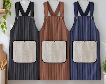 May include: Three aprons in black, brown, and navy blue, each featuring thin vertical white stripes, a large beige textured front pocket with black trim, and cross-back straps secured with metallic buttons at the chest.
