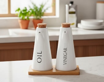 May include: Two white ceramic cruets with cork stoppers, one labeled OIL and the other VINEGAR in black lettering. The conical containers sit on a rectangular wooden tray. The design features a narrow spout at the top of each vessel.