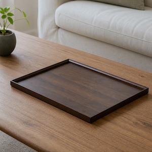 Puede incluir: Una bandeja de servicio rectangular hecha de madera de color marrón oscuro con una superficie plana y lisa y un borde elevado. La bandeja presenta un patrón de veta de madera visible en toda su estructura, mostrando un tono profundo y uniforme.