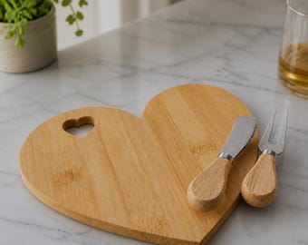 May include: A heart-shaped bamboo serving board featuring a small heart-shaped cutout near the top edge. The set includes a cheese spreader and a two-pronged cheese fork, both designed with matching light-toned wooden handles and stainless steel blades.