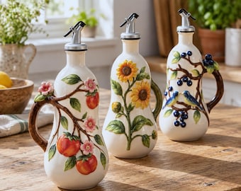May include: Three white ceramic oil dispensers with silver pour spouts and handles. The first features orange fruit and pink blossoms, the second displays yellow sunflowers, and the third shows blue birds perched on branches with dark berries.
