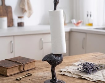 May include: A dark brown cast iron paper towel holder shaped like a standing bird with a long neck. The bird's neck serves as the vertical spindle, holding a single roll of white paper towels. The base is circular and supports the bird's two legs.