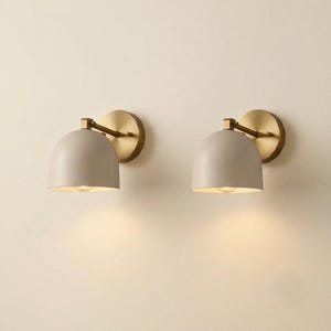May include: Two wall sconces featuring matte beige dome-shaped shades and brushed brass circular wall mounts. Each fixture has a short, adjustable arm connecting the shade to the base, emitting a warm, soft glow from the underside of the dome.