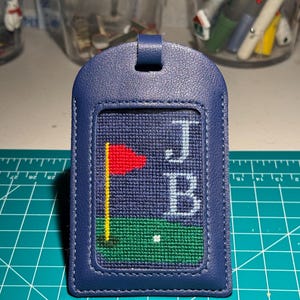 May include: A navy blue luggage tag with a stitched golf-themed design. The design includes a red flag, a green putting green, a white golf ball, and the initials "JB" in white. The tag has a loop at the top for attachment.